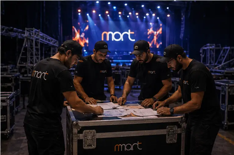 Equipe da NMont reunida em volta de um case, conferindo o roteiro com o palco montado ao fundo