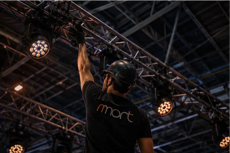 Técnico de capacete ajustando um refletor móvel preso à treliça de iluminação